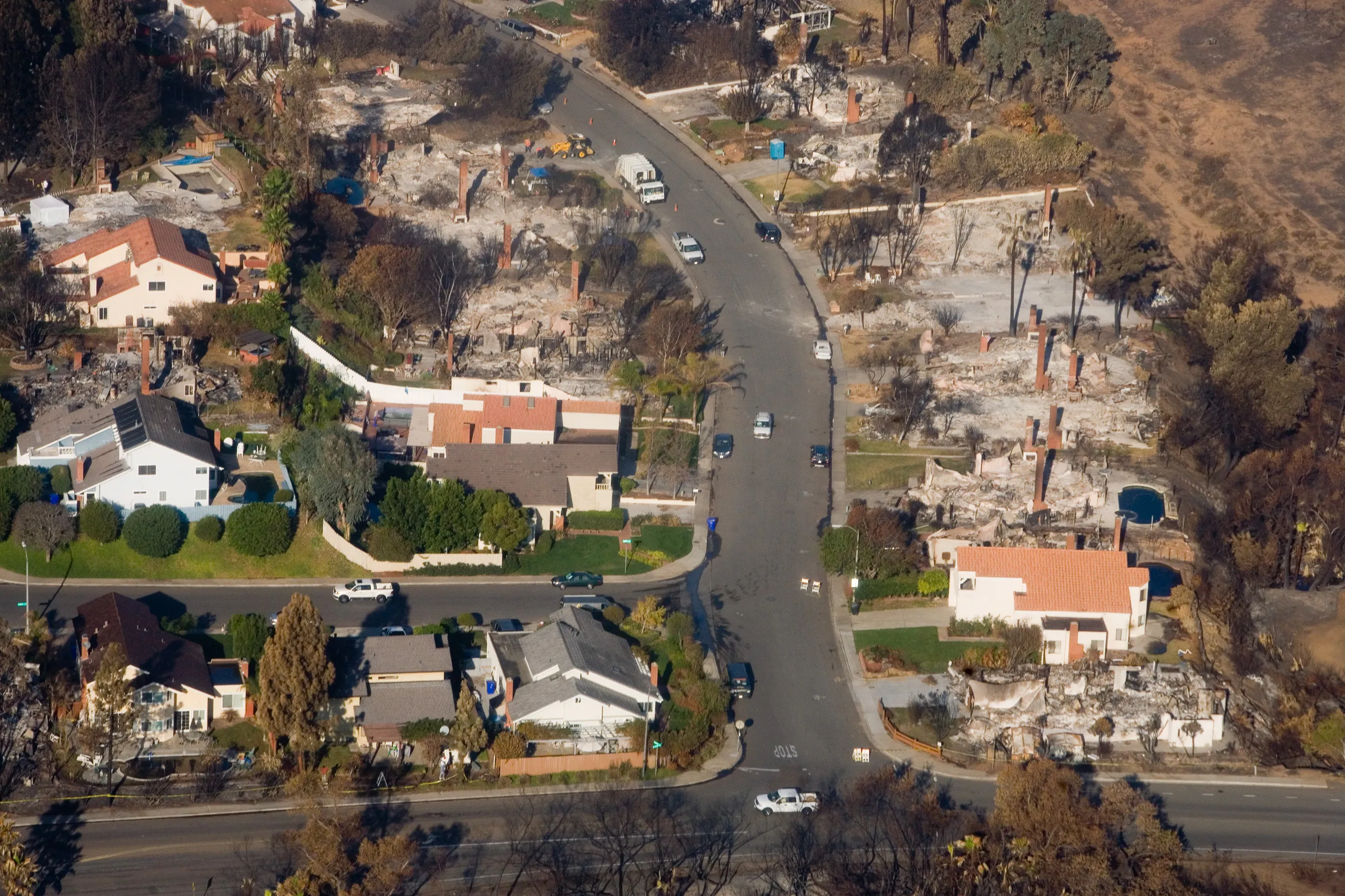 An aerial view of a California neighborhood reduced to ash and foundations by wildfire, with a few houses spared