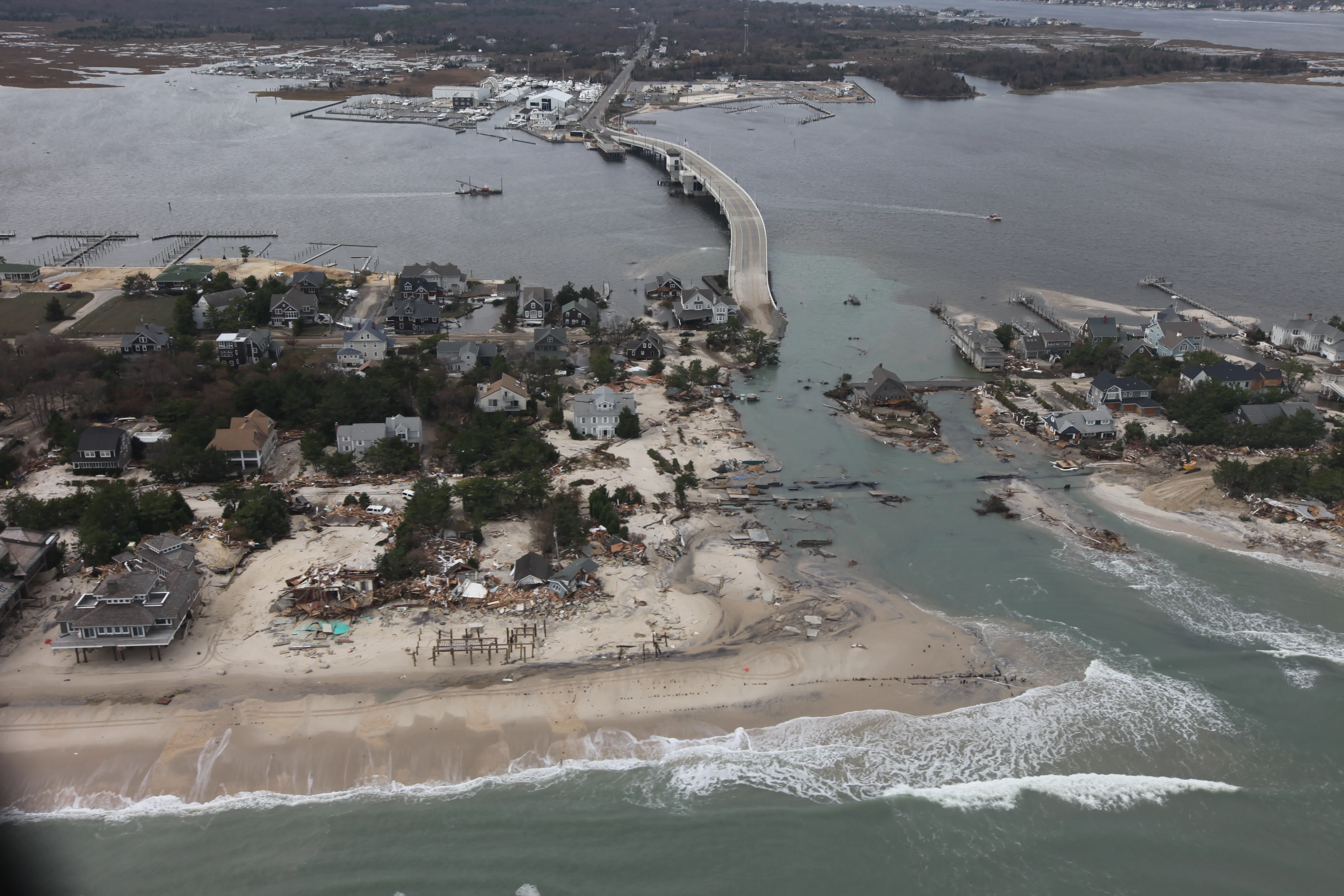 An aerial view of a barrier-island coastline breached by a hurricane, with a new inlet cut through destroyed beachfront homes