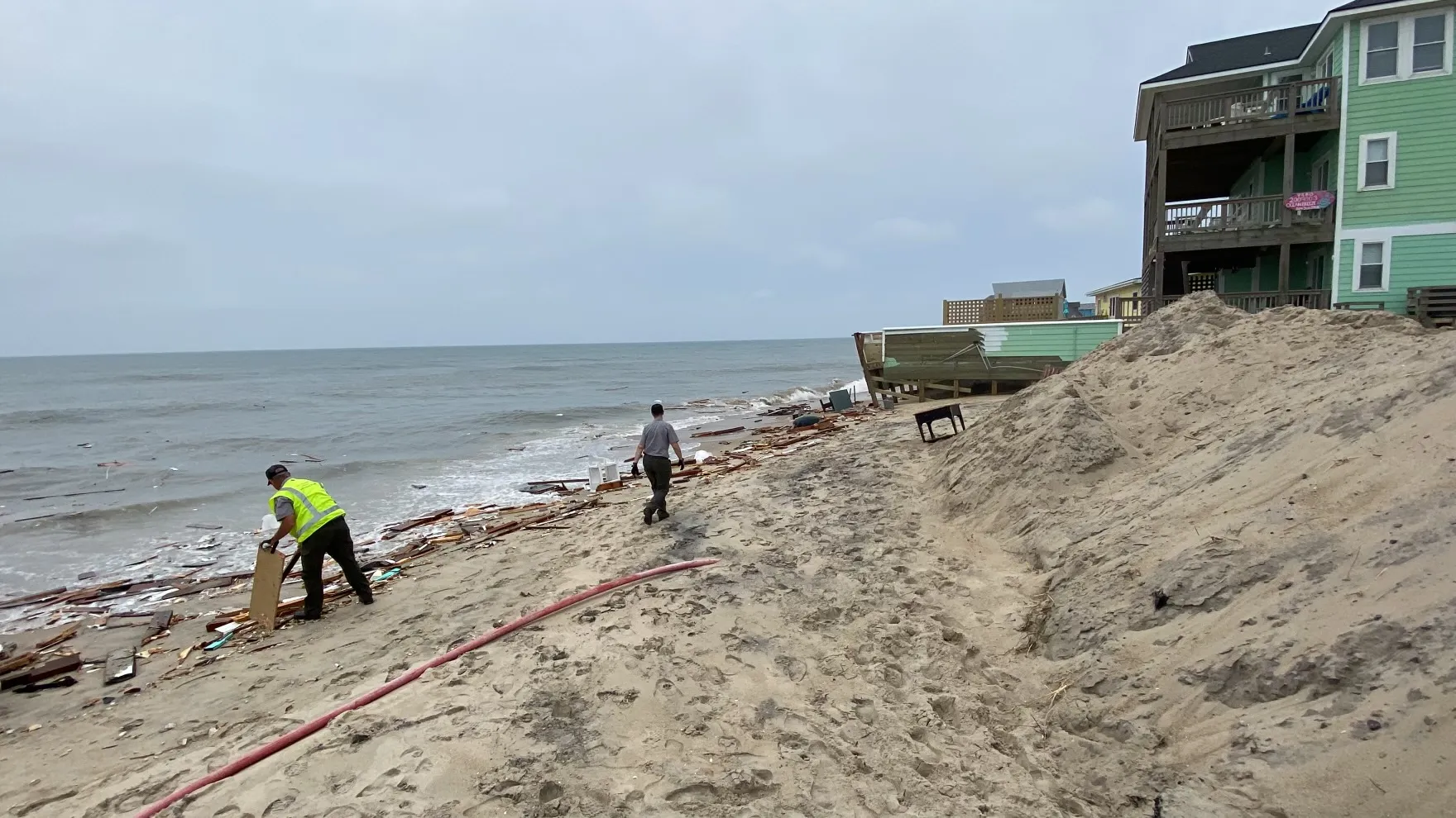 A beachfront house collapsed into debris on a severely eroded beach, with waves reaching the remaining homes