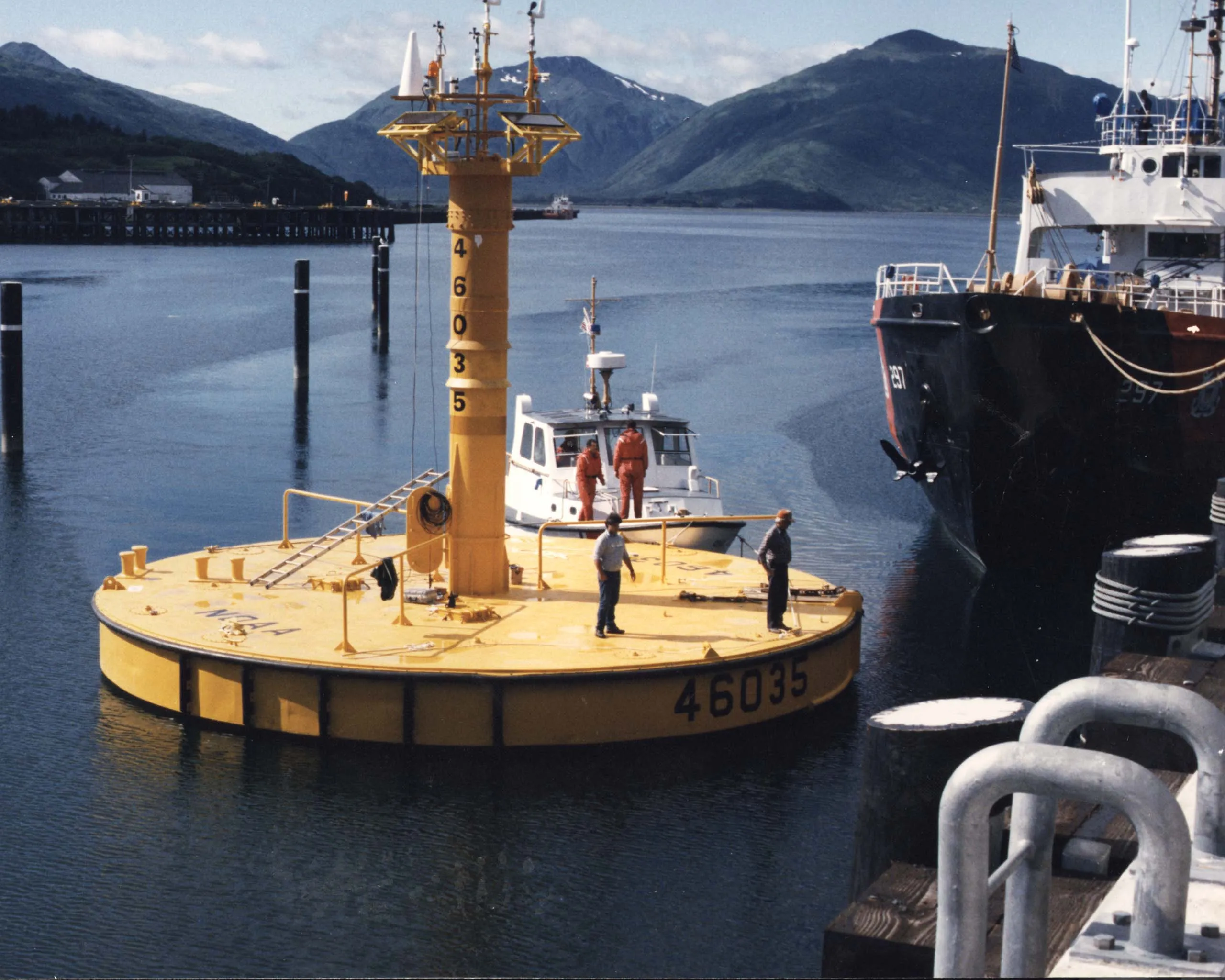 A large yellow NOAA weather buoy floating in a harbor next to a boat