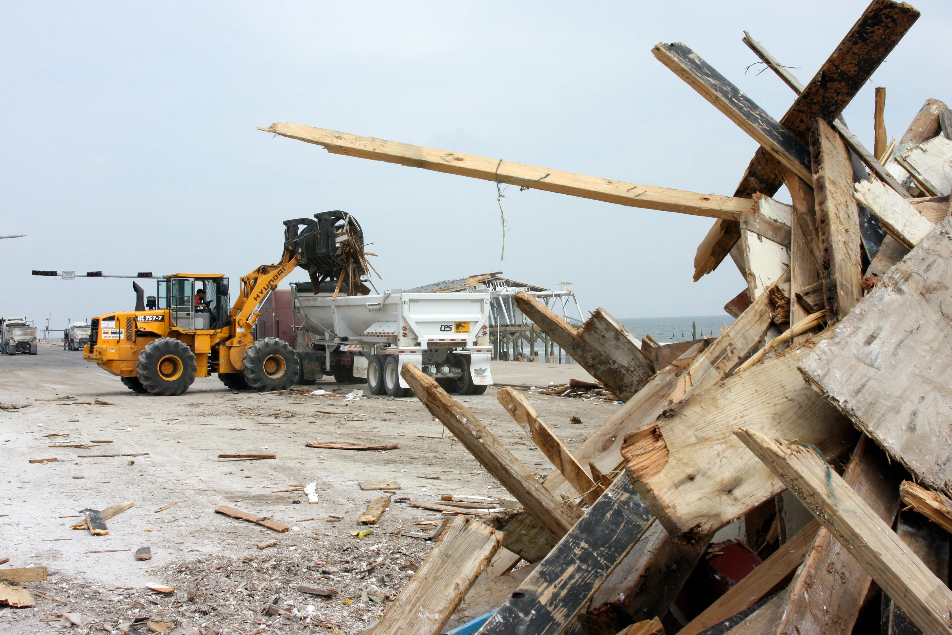 A front-end loader scooping a pile of hurricane debris into a dump truck on a residential street
