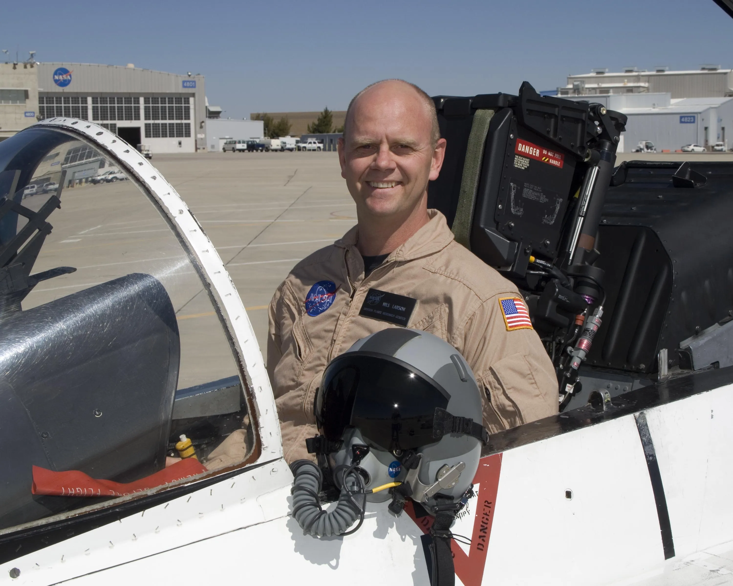A research pilot stands beside a jet cockpit holding a flight helmet with attached oxygen mask